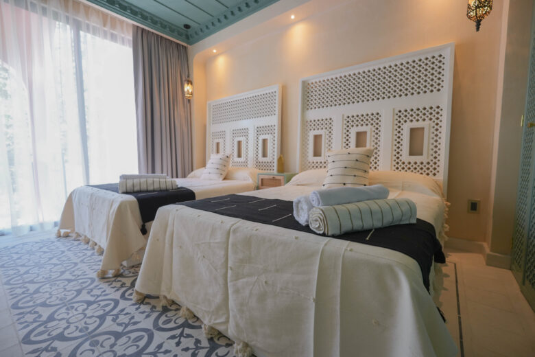 Habitación secundaria en una unidad de Pink Riad Tulum, con dos camas vestidas en textiles naturales, cabeceras talladas de estilo marroquí y luz natural filtrándose a través de cortinas ligeras. Espacio elegante de los departamentos en venta en Tulum.